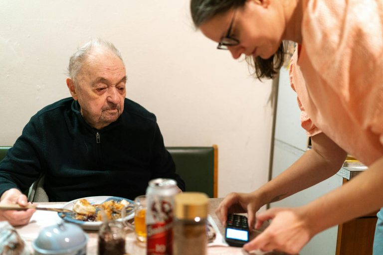 A caregiver assists an elderly man during mealtime at an indoor setting. Captured in Praha.