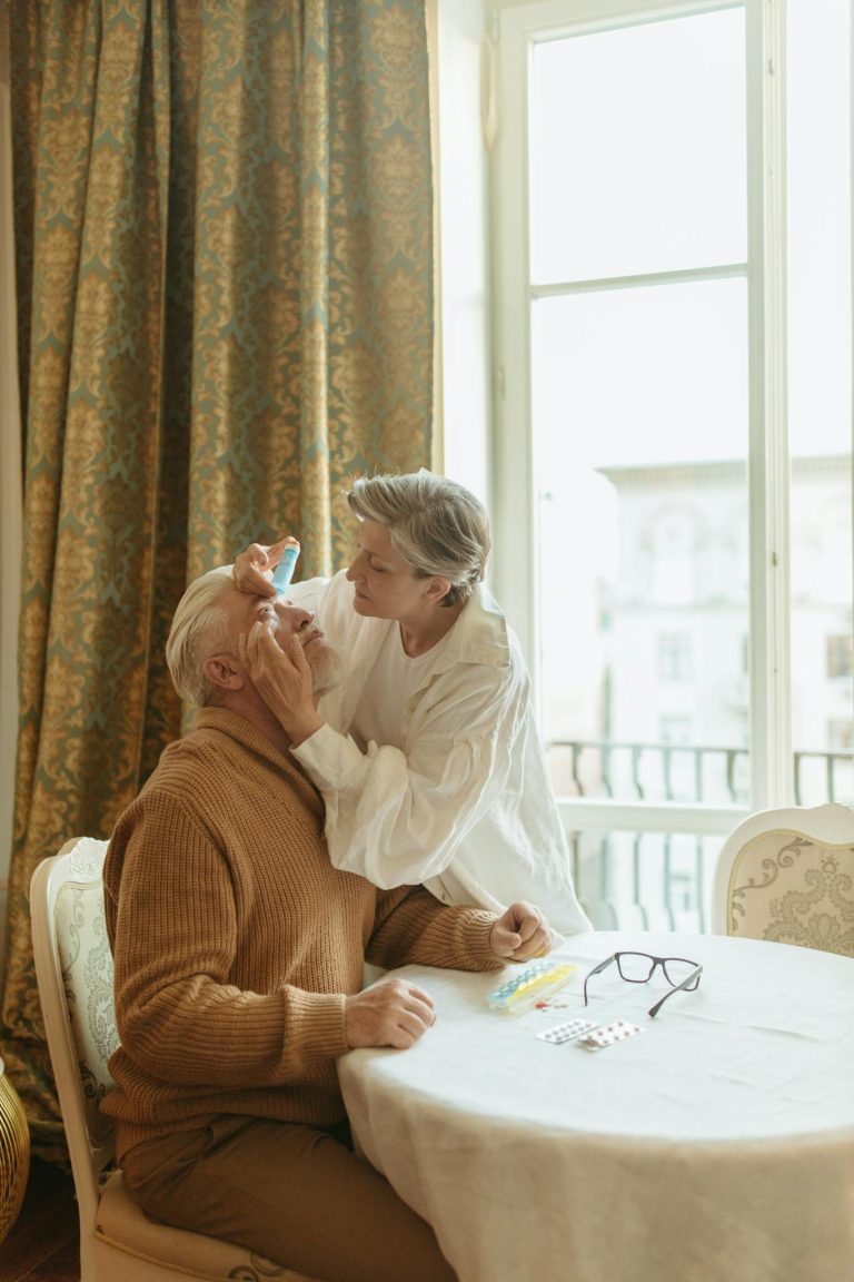 A senior woman assisting an elderly man with eye drops in a cozy indoor setting.